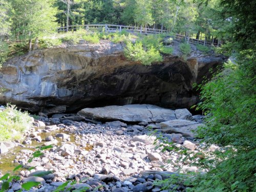 A rocky riverbed with large stones, surrounded by greenery and a wooden bridge above a natural rock formation.