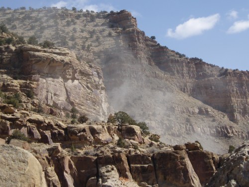 Dust rises from a rocky cliffside, showcasing rugged terrain and sparse vegetation under a blue sky with scattered clouds.