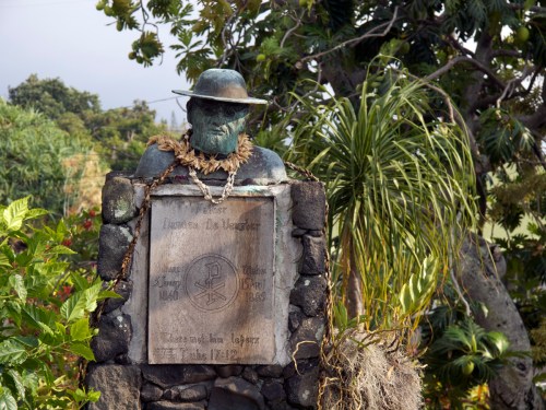 A bronze statue of a man wearing a hat, adorned with a lei, stands on a stone pedestal surrounded by greenery.