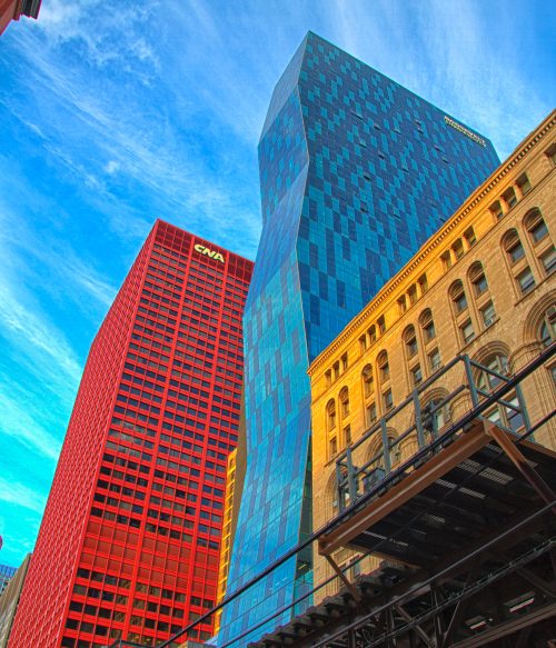 A vibrant cityscape featuring a red skyscraper, a blue modern building, and historic architecture under a blue sky.