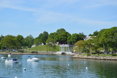 A calm waterfront scene with boats anchored, green trees, and a white-columned building in the background.