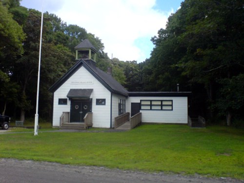 A white building with a bell tower, surrounded by trees, featuring a grassy area and a flagpole in front.