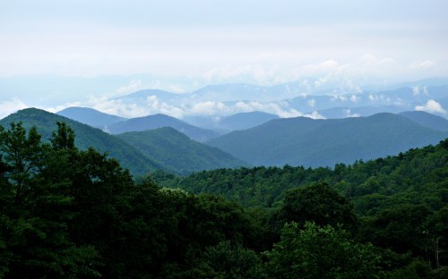 A panoramic view of lush green mountains shrouded in mist and clouds under a cloudy sky.