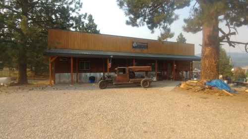 A rustic building with a wooden facade and a vintage car parked in front, surrounded by trees and gravel.