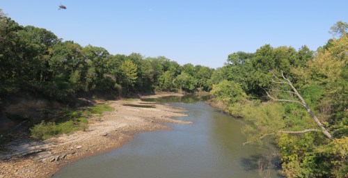 A serene river flows through a lush green landscape, with rocky banks and trees lining the water's edge.