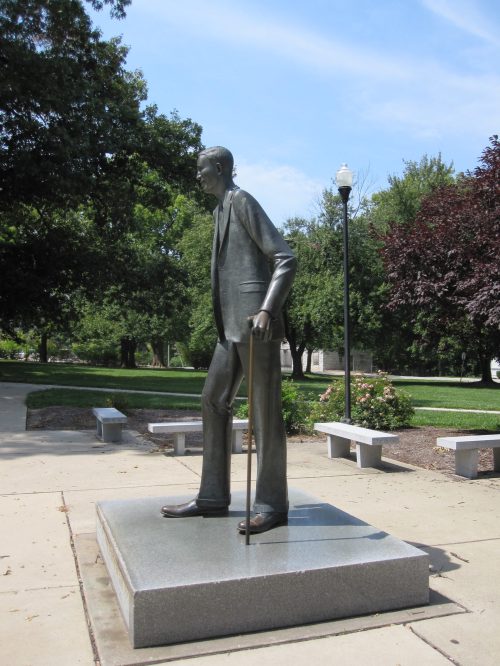 A bronze statue of a man in a suit, holding a cane, stands on a pedestal in a park setting.
