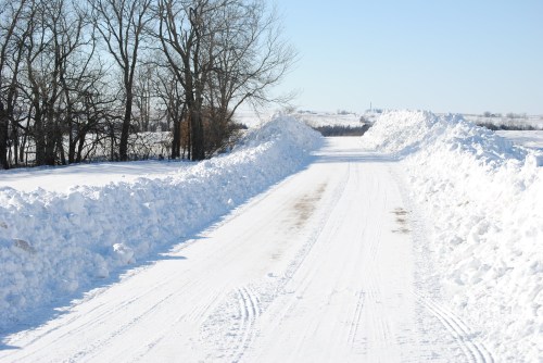 A snow-covered road flanked by tall snowbanks and bare trees under a clear blue sky.