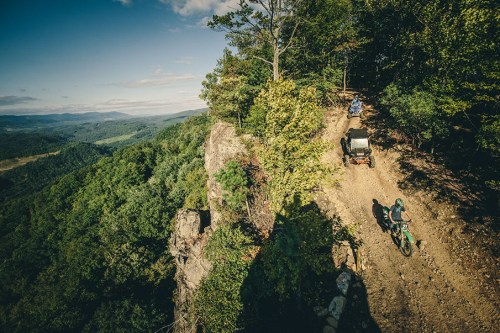 Aerial view of off-road vehicles navigating a rocky trail through lush green hills and trees.