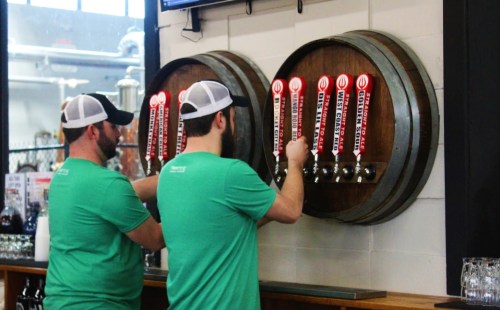 Two men in green shirts and white hats are pouring beer from taps mounted on wooden barrels.