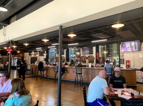 A lively bar interior with patrons seated at tables and the bar, featuring a TV and a variety of drinks on display.