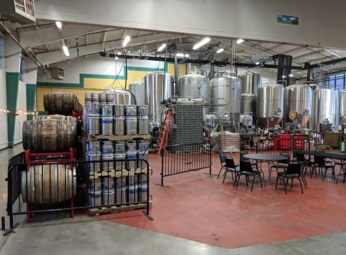 Interior of a brewery featuring large fermentation tanks, stacked barrels, and tables for seating.