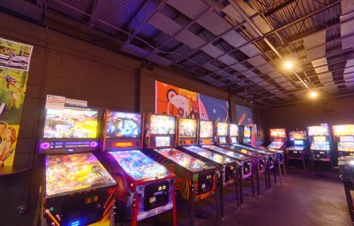 A row of colorful pinball machines in a dimly lit arcade, with vibrant artwork on the walls.