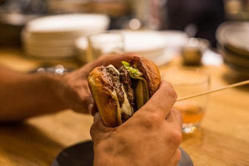 A person holds a juicy burger with lettuce and sauce, ready to take a bite, with plates and a drink in the background.