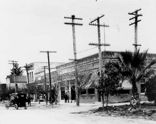 Historic black-and-white photo of a street with early 20th-century buildings, power poles, and a vintage car.