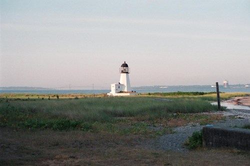 A white lighthouse stands on a grassy shore, with a bridge and distant buildings visible across the water.
