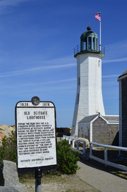 A historic lighthouse with a flag, next to a sign detailing its history, surrounded by a coastal landscape.
