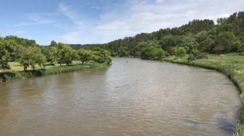 A serene river curves through lush greenery under a clear blue sky, surrounded by trees and grassy banks.