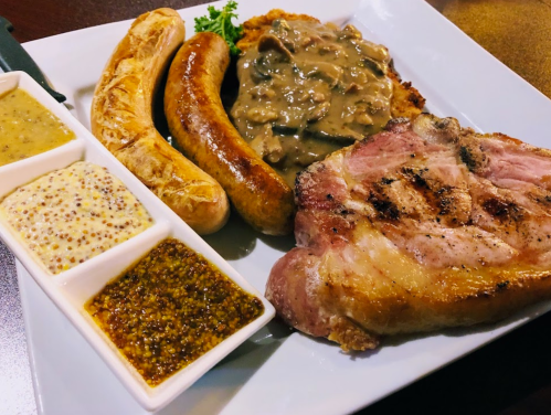 A plate featuring grilled meats, including sausages and a pork chop, accompanied by various mustards and a creamy sauce.