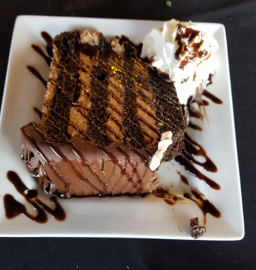 A slice of chocolate cake topped with whipped cream and drizzled with chocolate sauce on a white plate.