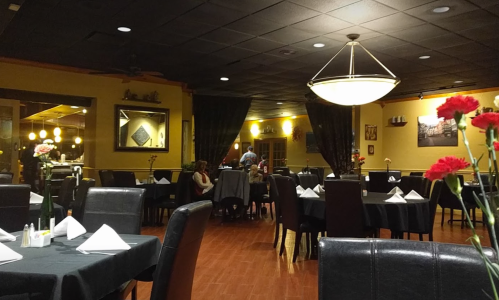 A cozy restaurant interior with dark tables, white napkins, and flowers, featuring warm lighting and a welcoming atmosphere.