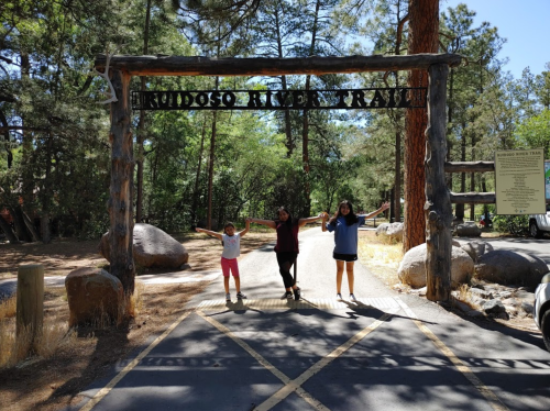 Three children stand under a wooden archway labeled "Ruidoso River Trail," surrounded by trees and rocks.
