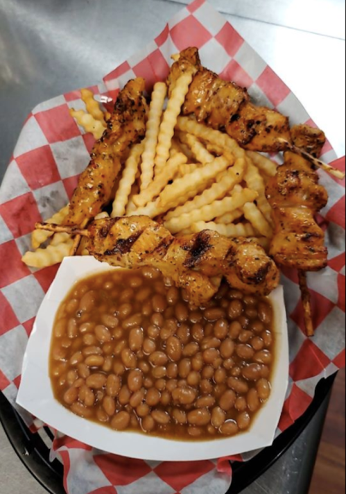 A plate with grilled chicken skewers, crinkle-cut fries, and a serving of baked beans on a checkered paper.