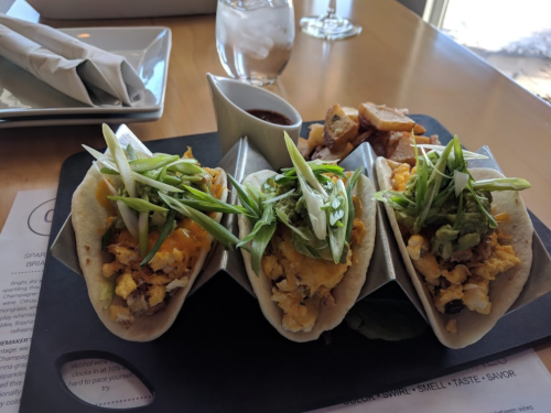 Three tacos filled with scrambled eggs, cheese, and green toppings, served with a side of sauce and crispy potatoes.