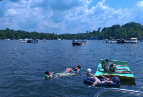 People relaxing on floating mats in a lake, surrounded by boats and lush greenery under a blue sky with clouds.