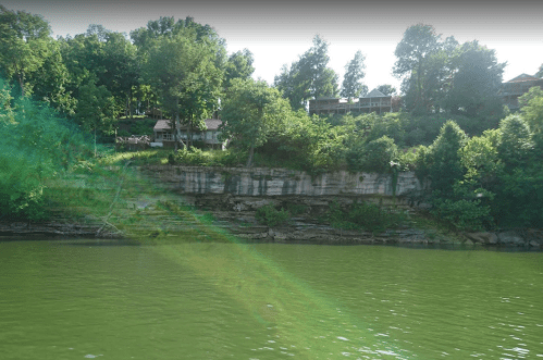 Lush green trees and a rocky shoreline beside a calm, green lake with houses in the background.