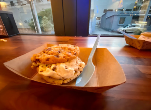 A cookie sandwich with chocolate chip cookies and ice cream, served in a paper tray on a wooden table.