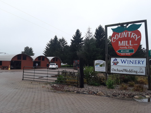 Sign for Country Mill with a welcoming message, surrounded by trees and buildings, on a cloudy day.