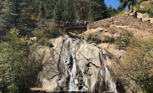 A waterfall cascades over rocks, with a wooden bridge and people in the background surrounded by trees.