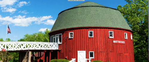 A red, round barn with a green roof and white trim, featuring a sign that says "MATTRESSES" and an American flag nearby.