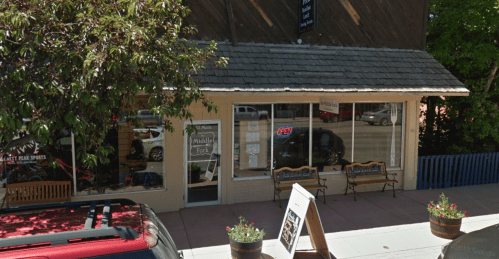 A storefront with an "Open" sign, benches outside, and potted plants, set in a tree-lined street.