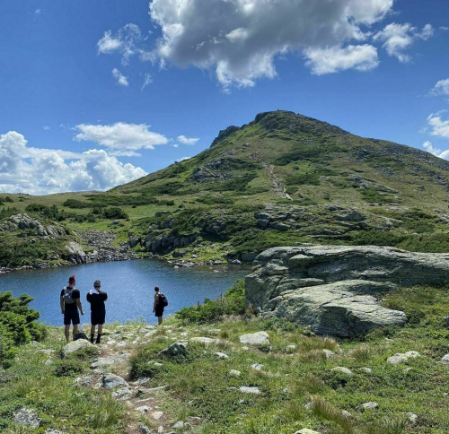 Three hikers stand by a serene lake, surrounded by lush greenery and a mountain under a bright blue sky.