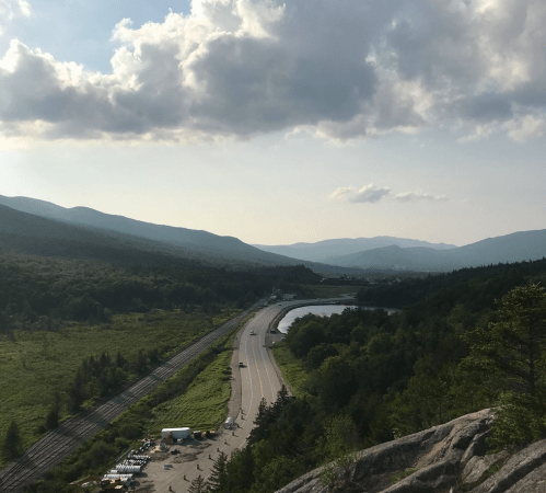 A scenic view of a winding road and river surrounded by mountains and lush greenery under a partly cloudy sky.