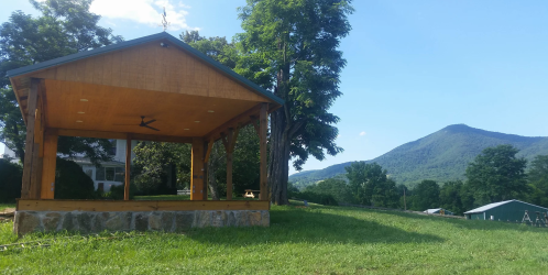 A wooden pavilion on a grassy area with mountains in the background under a clear blue sky.
