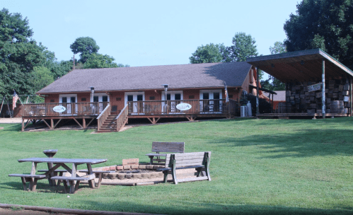 A rustic building with a porch and picnic area, surrounded by green grass and trees on a sunny day.