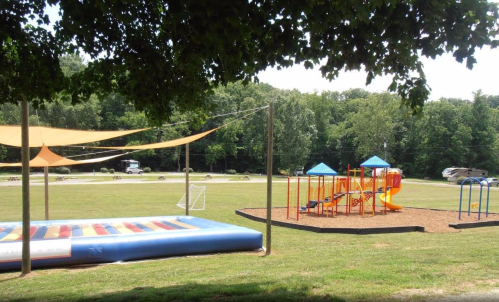 A playground with colorful equipment, a large inflatable bounce area, and shaded seating in a grassy park.