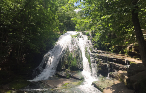 A serene waterfall cascading over rocks, surrounded by lush green trees and a bright blue sky.
