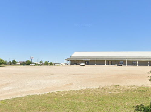 A large, empty parking lot in front of a single-story building under a clear blue sky.