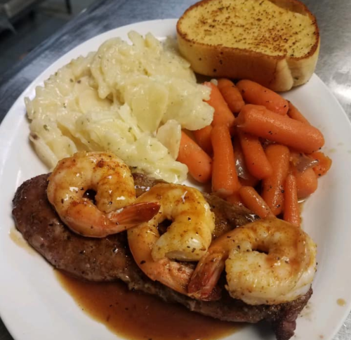 A plate featuring shrimp on steak, macaroni and cheese, glazed carrots, and a slice of garlic bread.