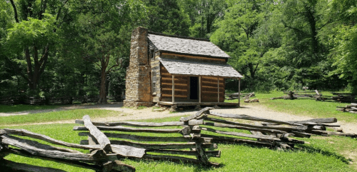 A rustic log cabin with a stone chimney, surrounded by greenery and a wooden fence in a peaceful outdoor setting.