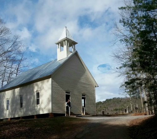 A small, white church with a steeple, surrounded by trees and a cloudy sky. A person stands at the entrance.