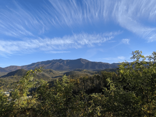 A panoramic view of mountains under a blue sky with wispy clouds, surrounded by lush greenery.