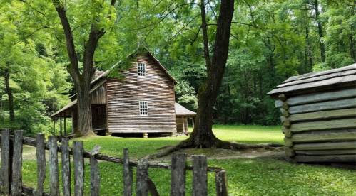 A rustic wooden house surrounded by lush green trees and a small log cabin nearby, with a wooden fence in the foreground.