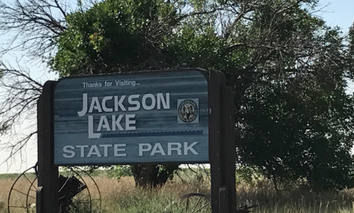 Sign for Jackson Lake State Park, featuring a blue background and surrounded by greenery.