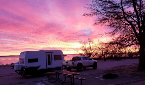 A camper and truck parked by a lake at sunset, with vibrant pink and purple skies and bare trees in the background.