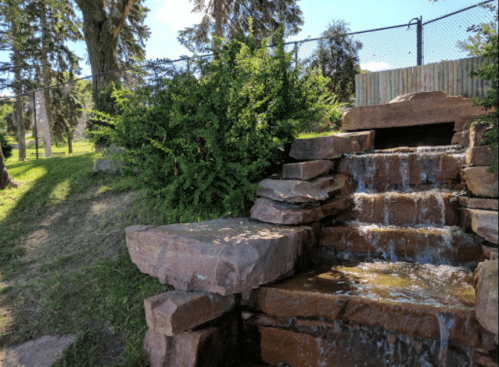 A small stone waterfall flows over rocks, surrounded by greenery and a fence in a sunny park setting.