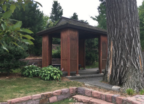 A wooden gazebo surrounded by greenery and trees, with a stone pathway leading through a garden.